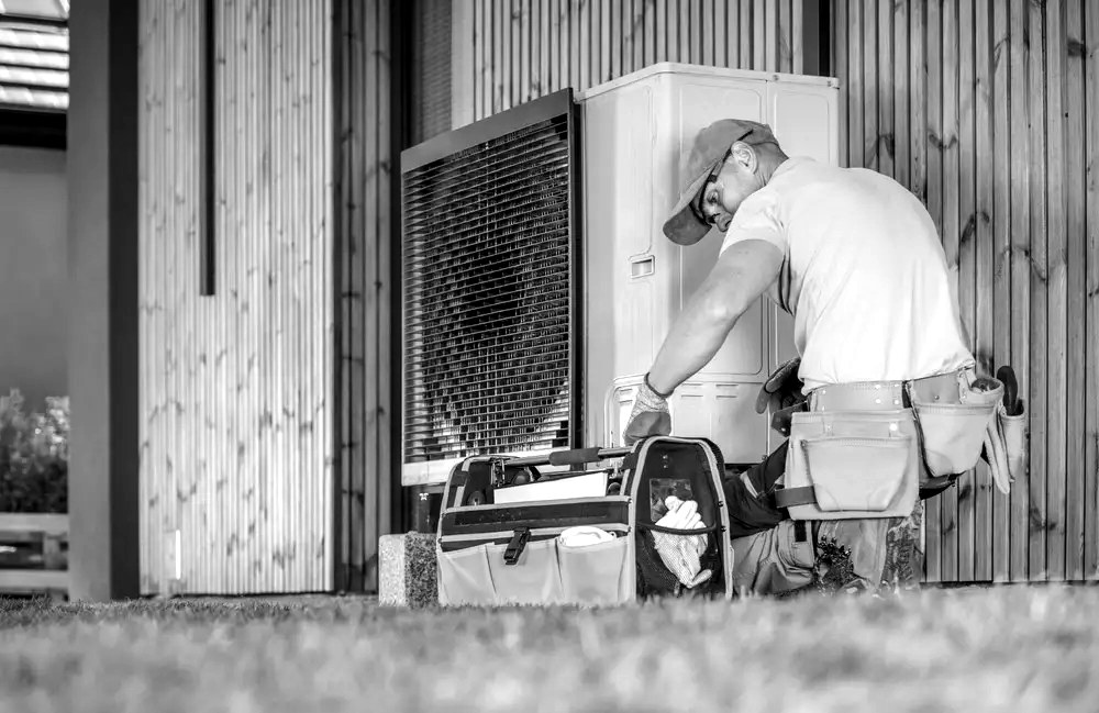 Black and white photograph of a technician kneeling to service an outdoor heat pump unit